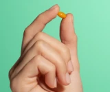 Hand holding a small orange pill against a green background.