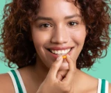 Smiling woman holding a small yellow-orange capsule near her mouth.