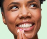 Person smiling while holding a small pink pill near their lips.