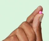 Fingers holding a small pink pill against a light green background.