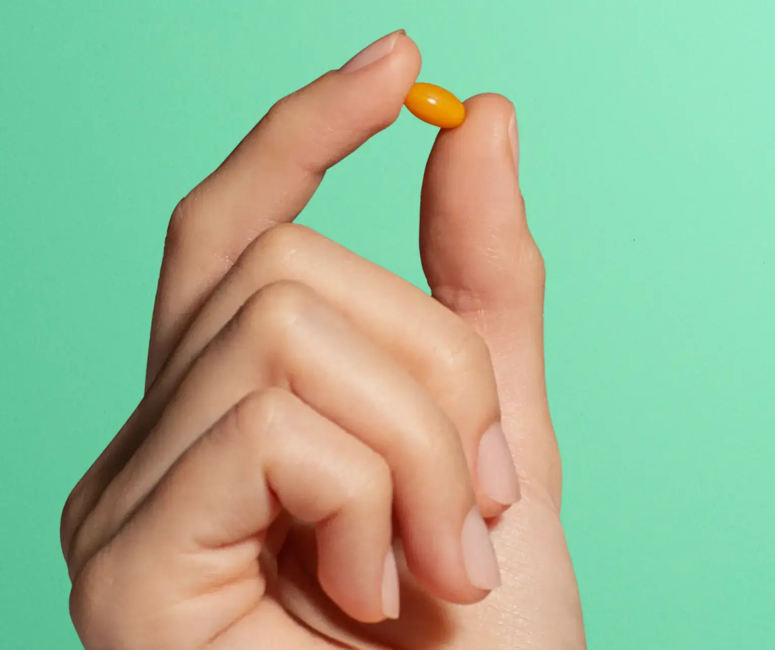 Hand holding a small orange pill against a green background.