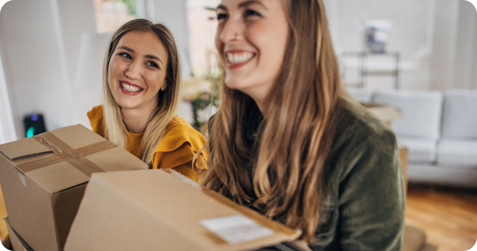 Two women smiling while holding cardboard boxes indoors.