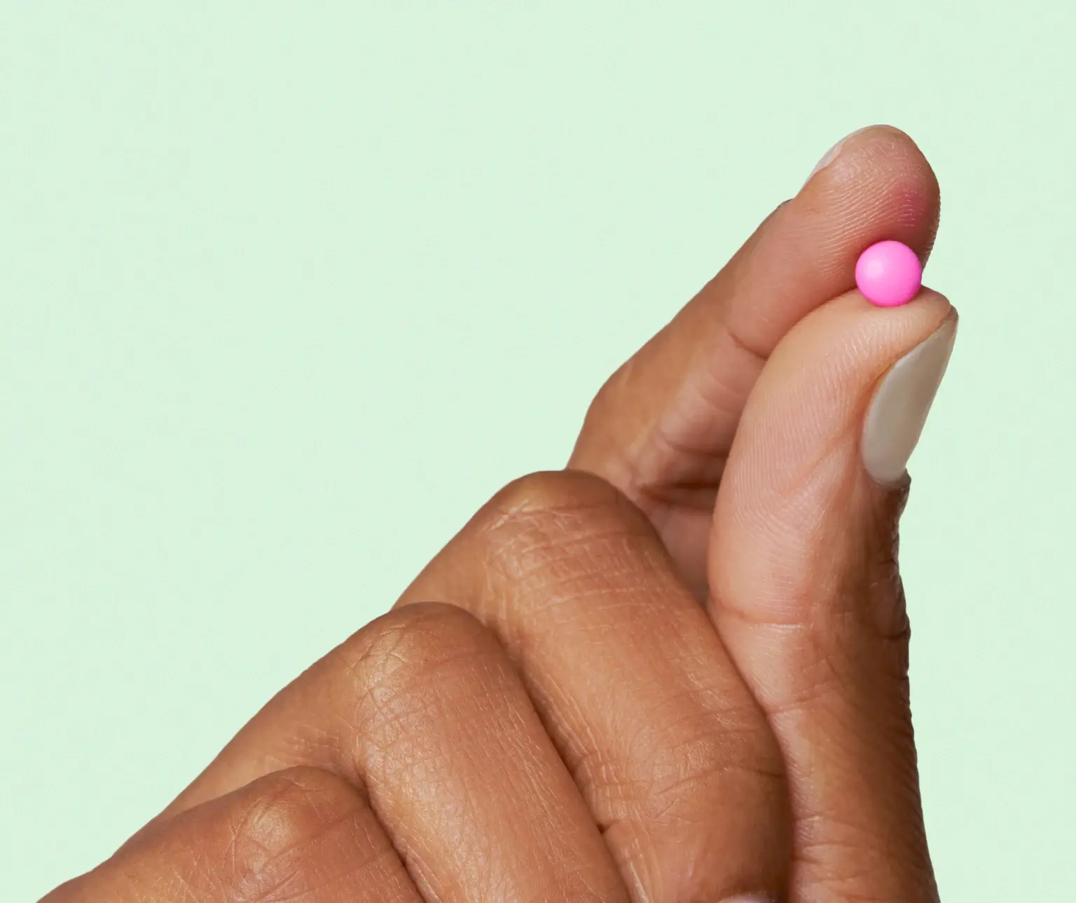 Fingers holding a small pink pill against a light green background.