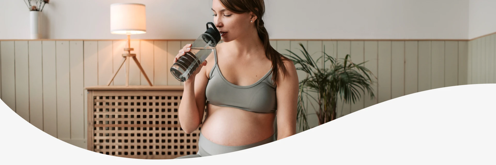 Pregnant woman in activewear drinking water from a bottle indoors.