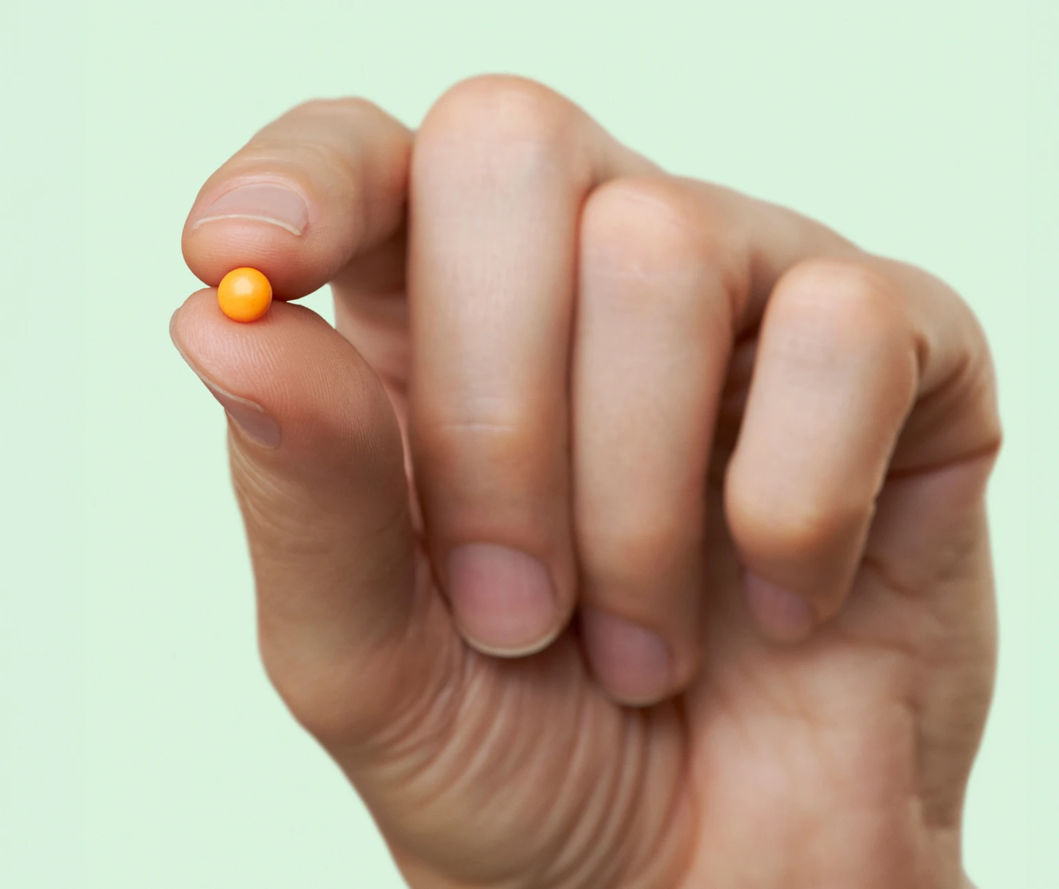 A hand holds a small orange bead between fingertips against a light green background.
